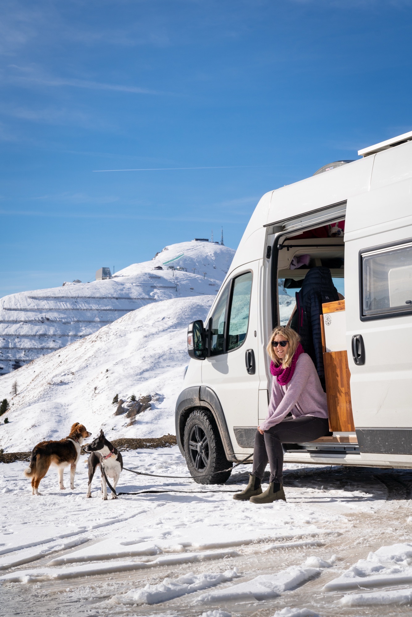 Woman sitting in van doorway with dogs in a snowy mountain setting